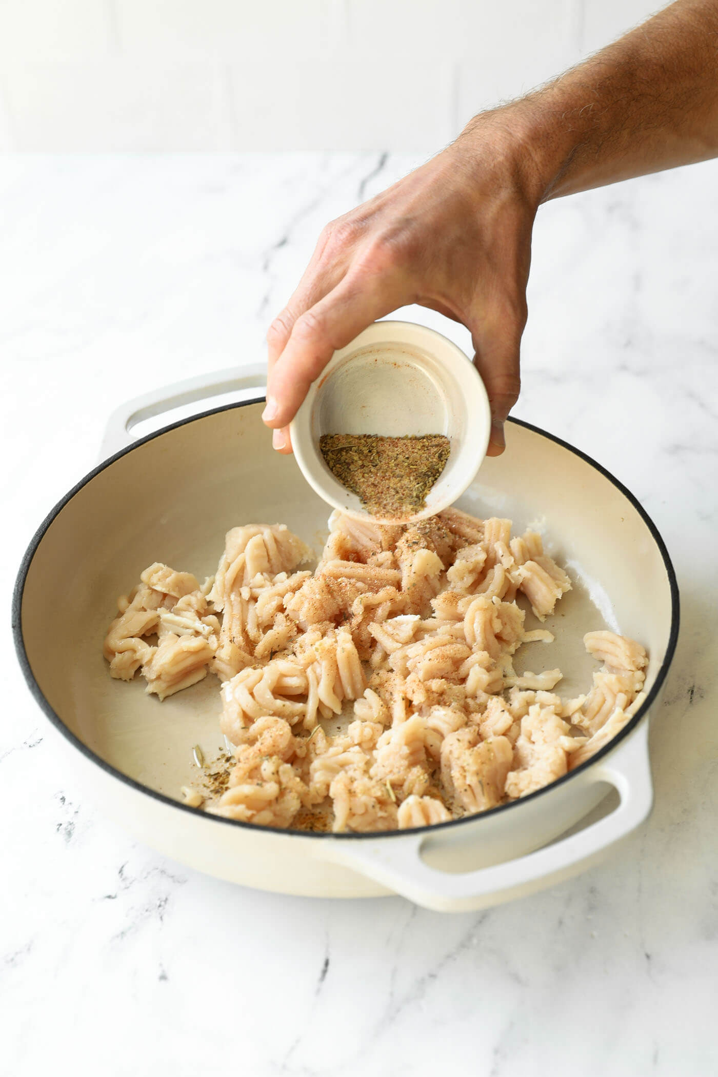 A hand pours seasoning from a small bowl onto chopped seafood in a white pan on a marble countertop.