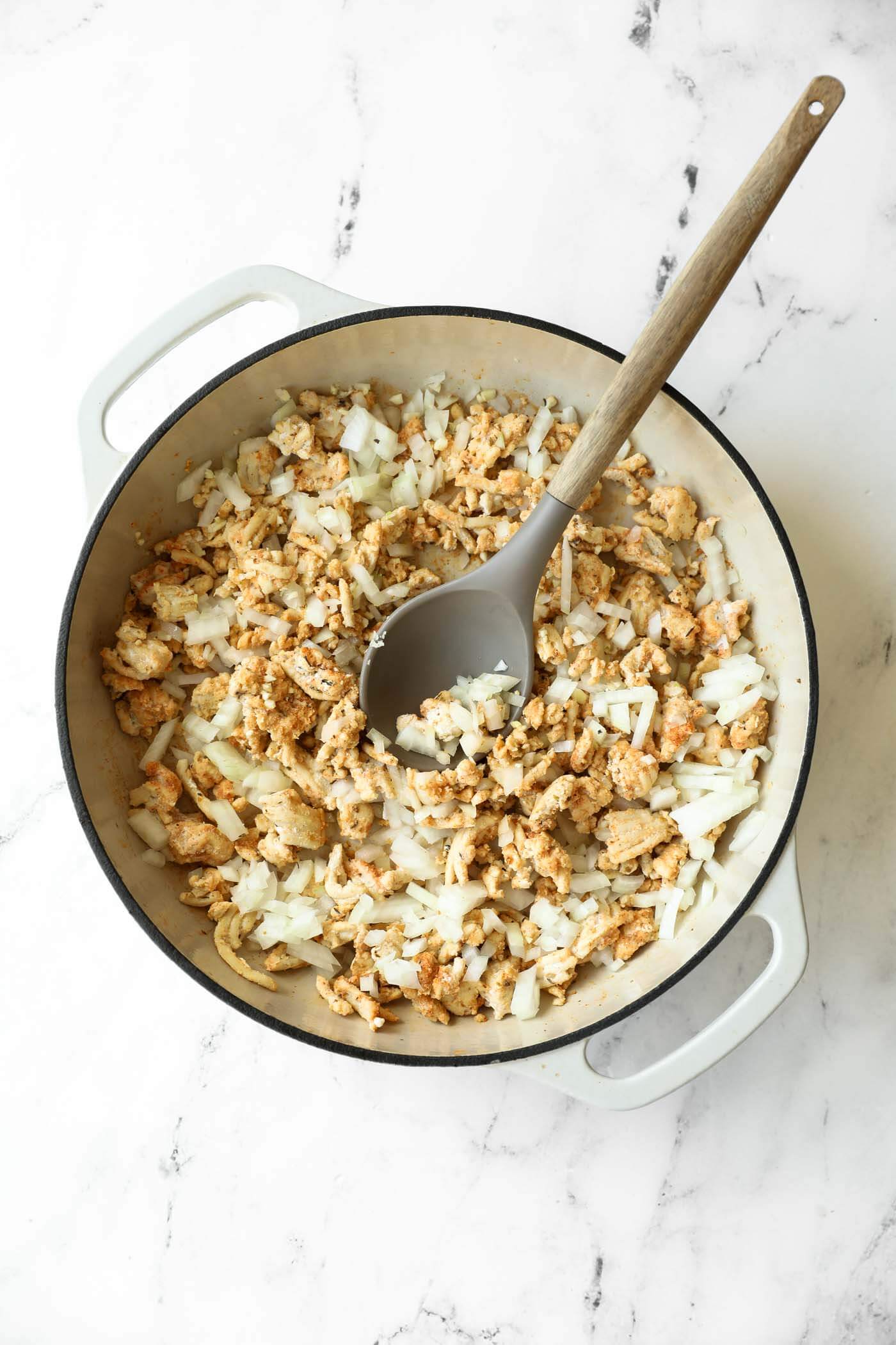 A white pot with cooked ground meat and chopped onions being stirred with a gray spoon on a marble countertop.