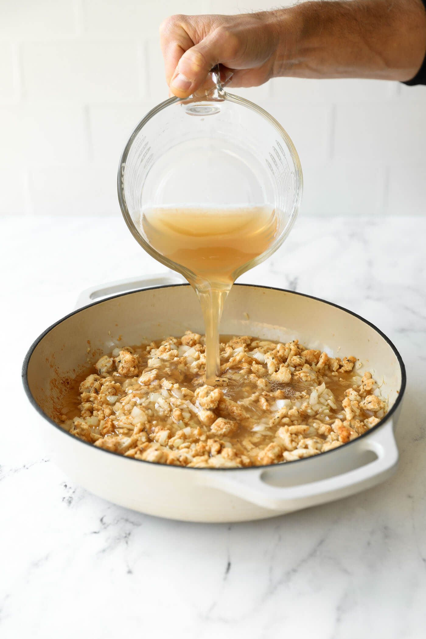 A hand pours broth from a glass measuring cup into a white skillet containing cooked ground meat and onions on a marble countertop.