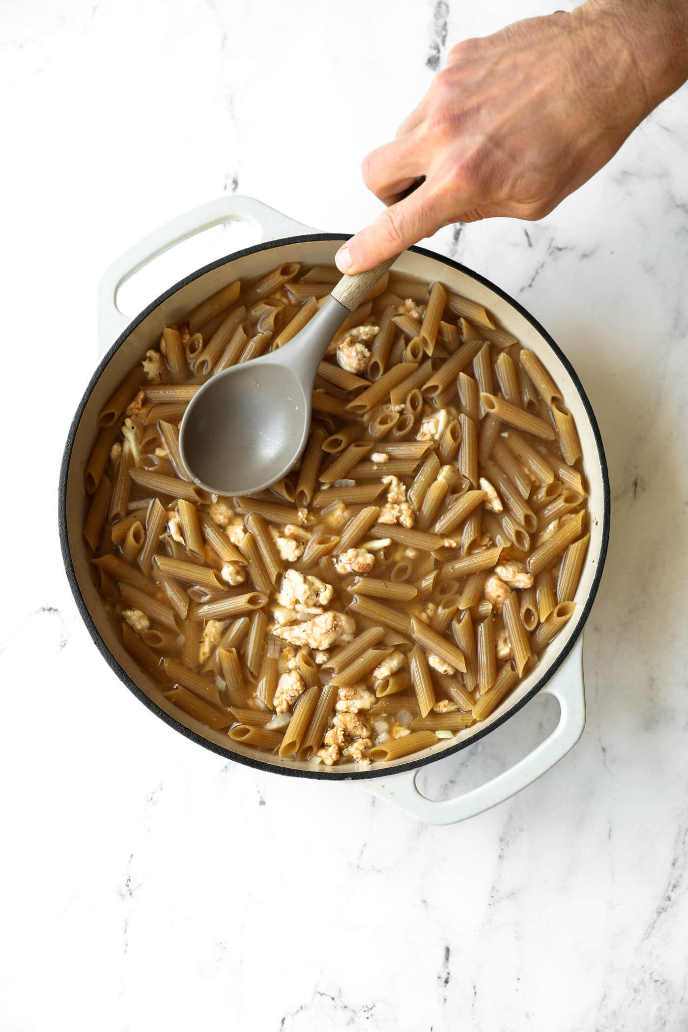 A hand stirs uncooked penne pasta and ground meat in a pot filled with water on a marble countertop.
