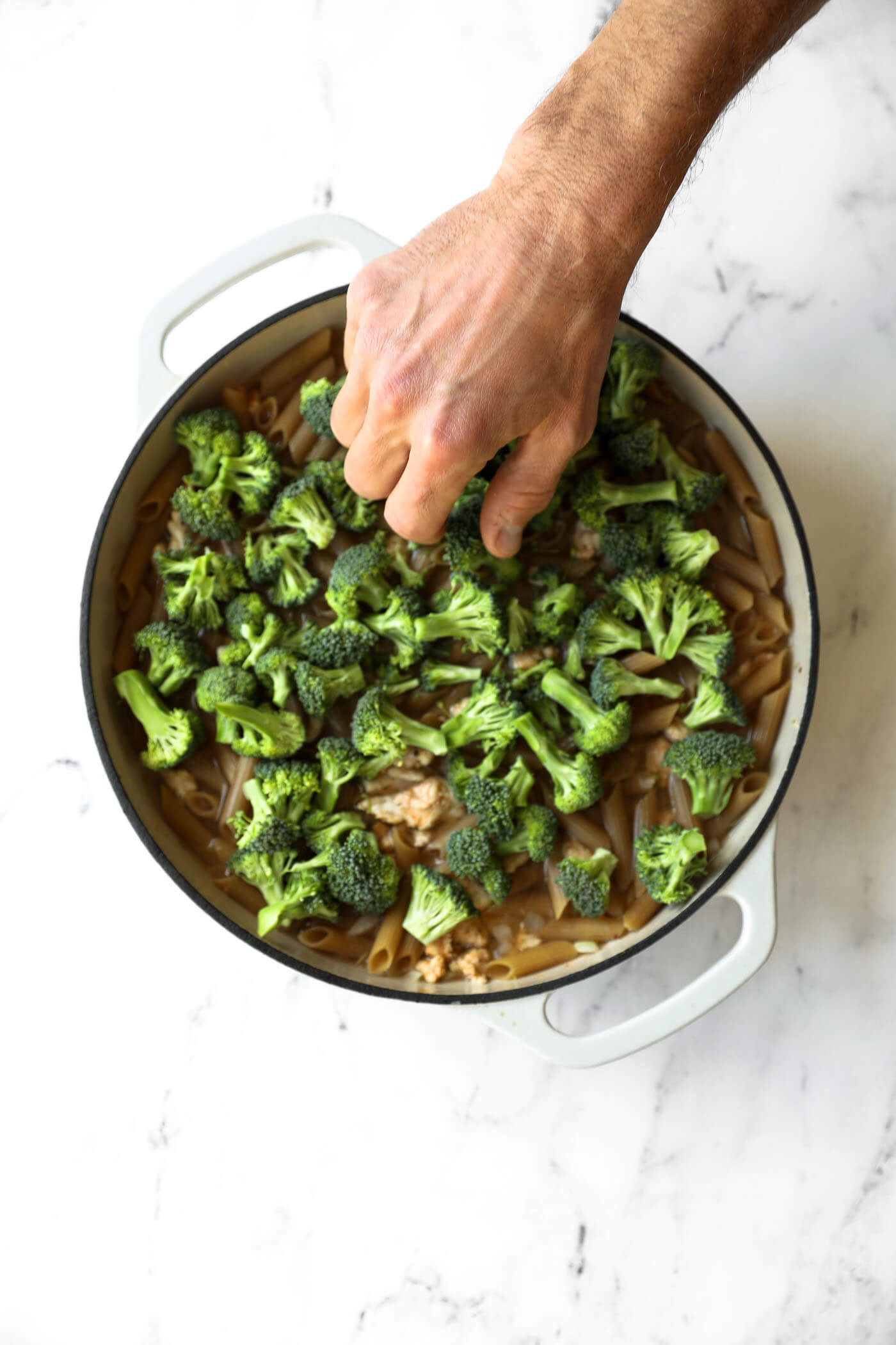 A hand adds broccoli florets to a pot of uncooked pasta and ground meat on a white marble countertop.