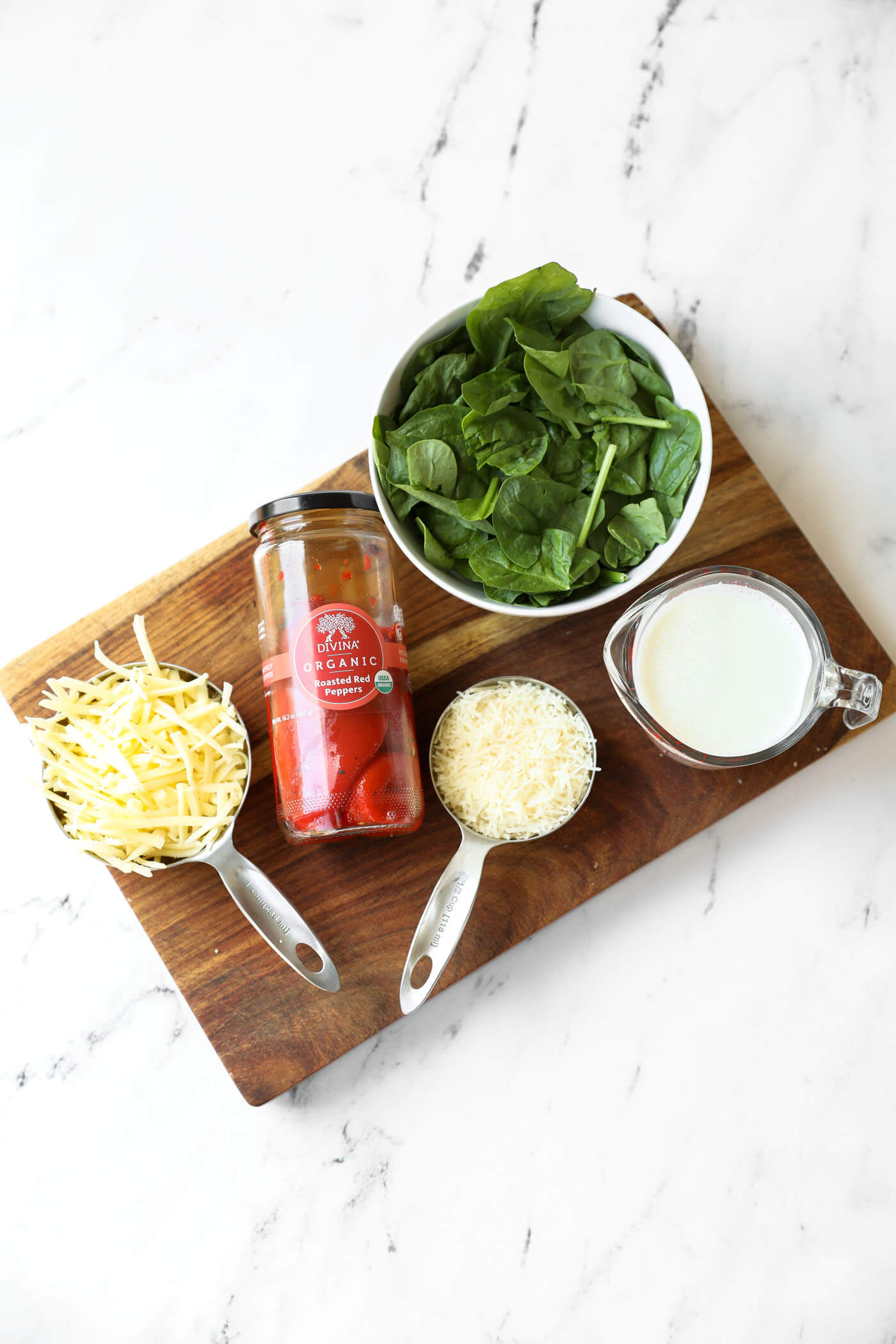 A bowl of spinach, a jar of roasted red peppers, shredded cheese, grated cheese, and a measuring cup of milk on a wooden board.
