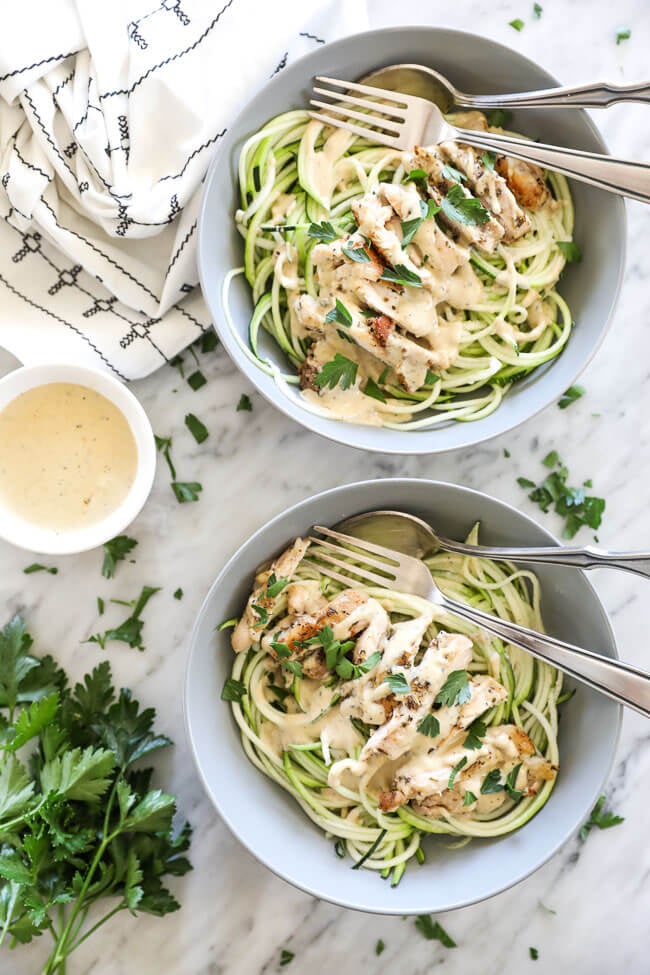 Overhead vertical image of two bowls of creamy chicken alfredo with a fork and spoon in each bowl. Extra sauce on the side with chopped parsley spread about. 