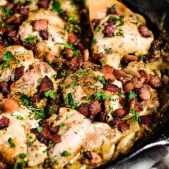 Close up angle image of creamy garlic chicken thighs in a cast iron skillet with chopped bacon and parsley on top