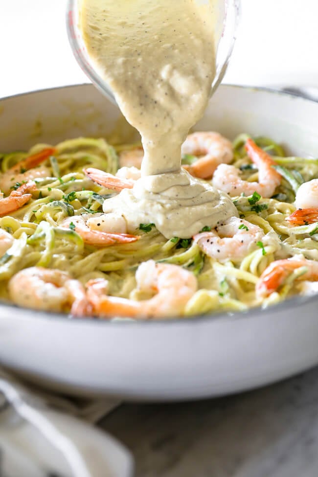 Angled image of pouring creamy sauce over skillet of zucchini noodles and shrimp. 