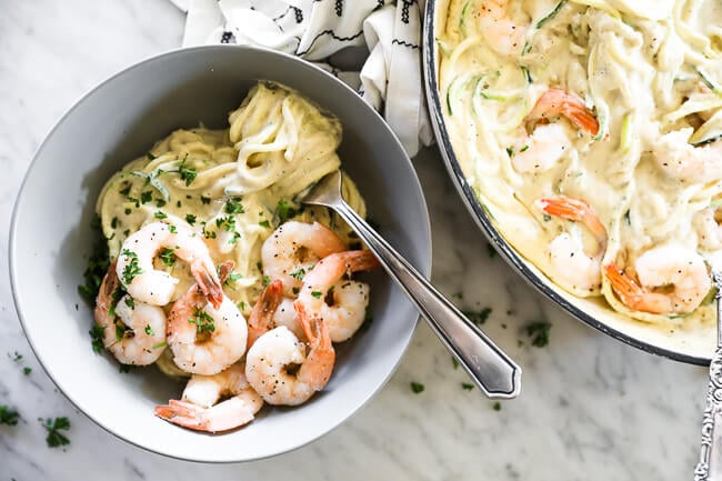 Horizontal overhead image of creamy garlic shrimp with zoodles served in a bowl with a fork with noodles wrapped around it. Skillet of dish off to the side and parsley sprinkled on top. 