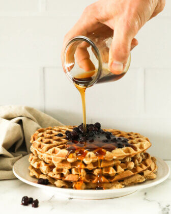 Image of a hand pouring maple syrup on a stack of waffles with wild blueberries on top.