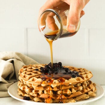 Image of a hand pouring maple syrup on a stack of waffles with wild blueberries on top.