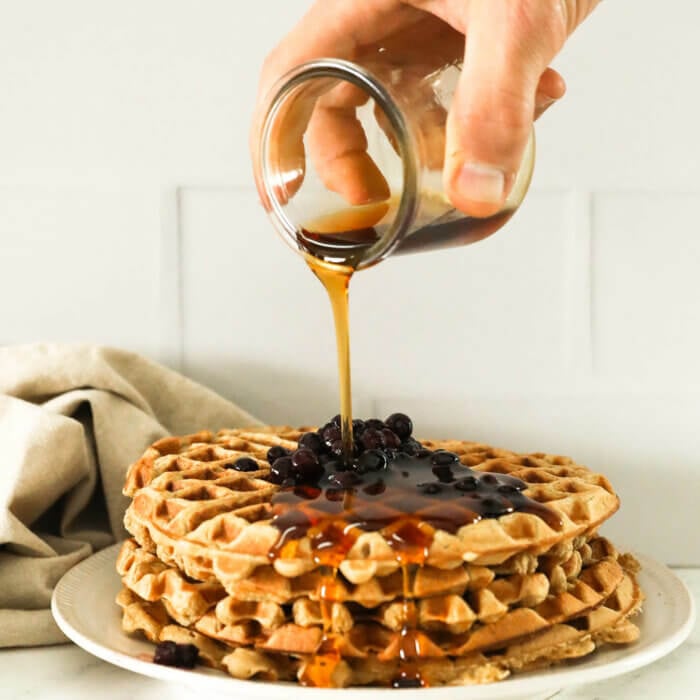 Image of a hand pouring maple syrup on a stack of waffles with wild blueberries on top.