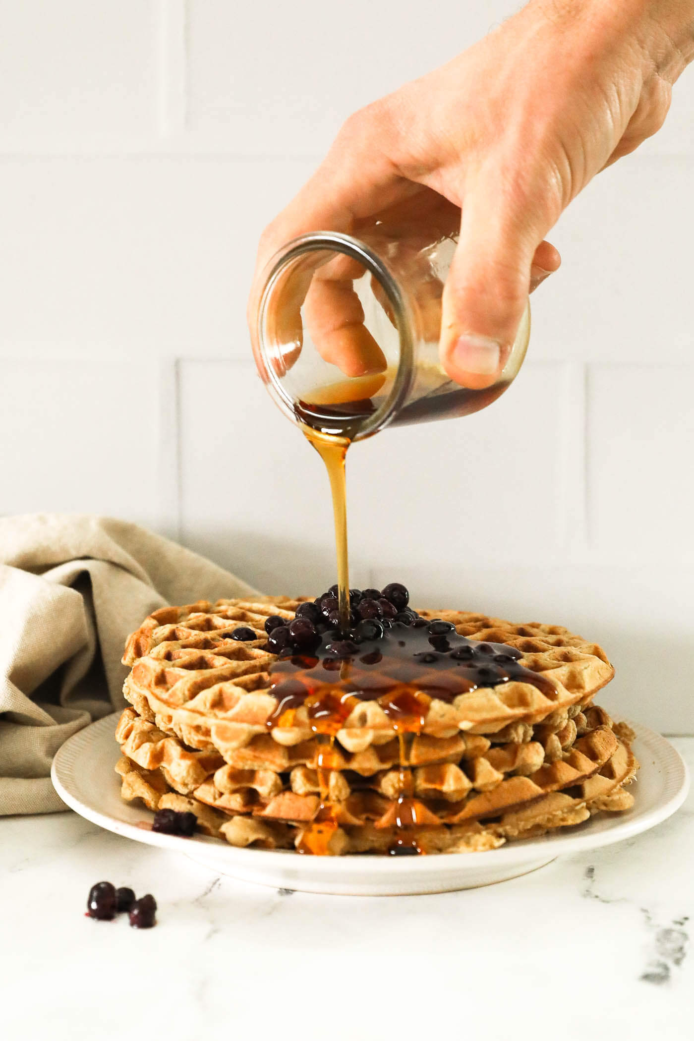 Image of a hand pouring maple syrup on a stack of waffles with wild blueberries on top.