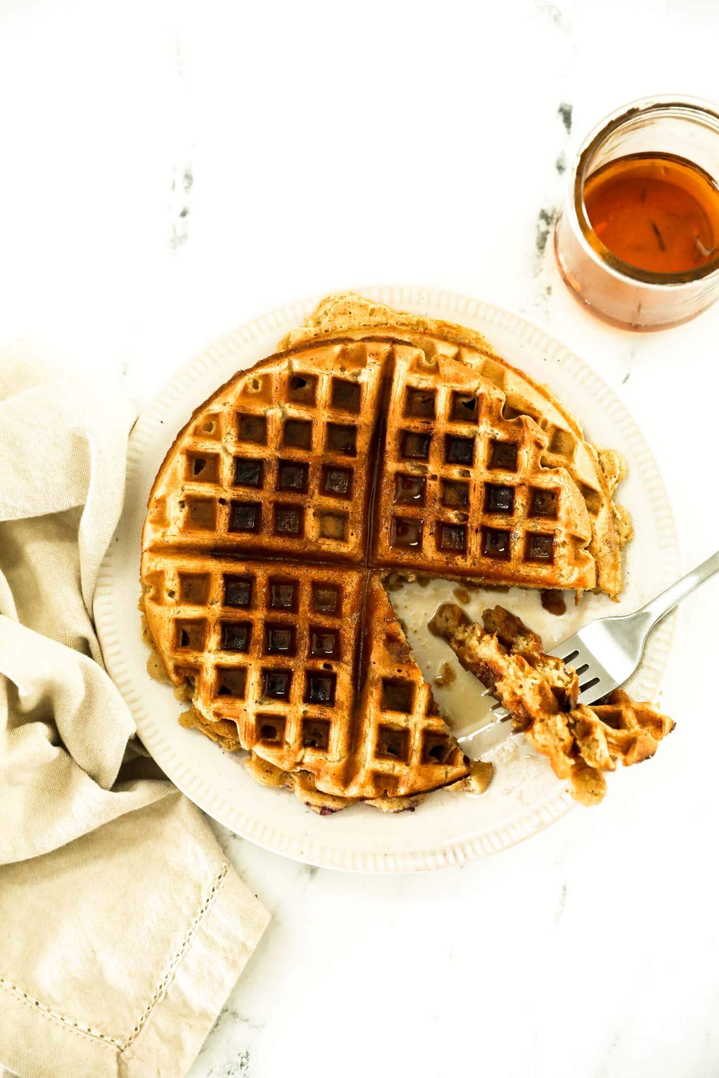 A plate with oat flour waffles. A triangle bite has been cut out and is on the fork next to the rest o the waffle. Maple syrup has been poured on top and more is on the side.