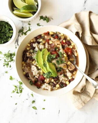 A bowl of vegetable soup with rice, beans, corn, tomato, and sliced avocado, garnished with cilantro, sits next to lime wedges and additional chopped cilantro on a marble surface.
