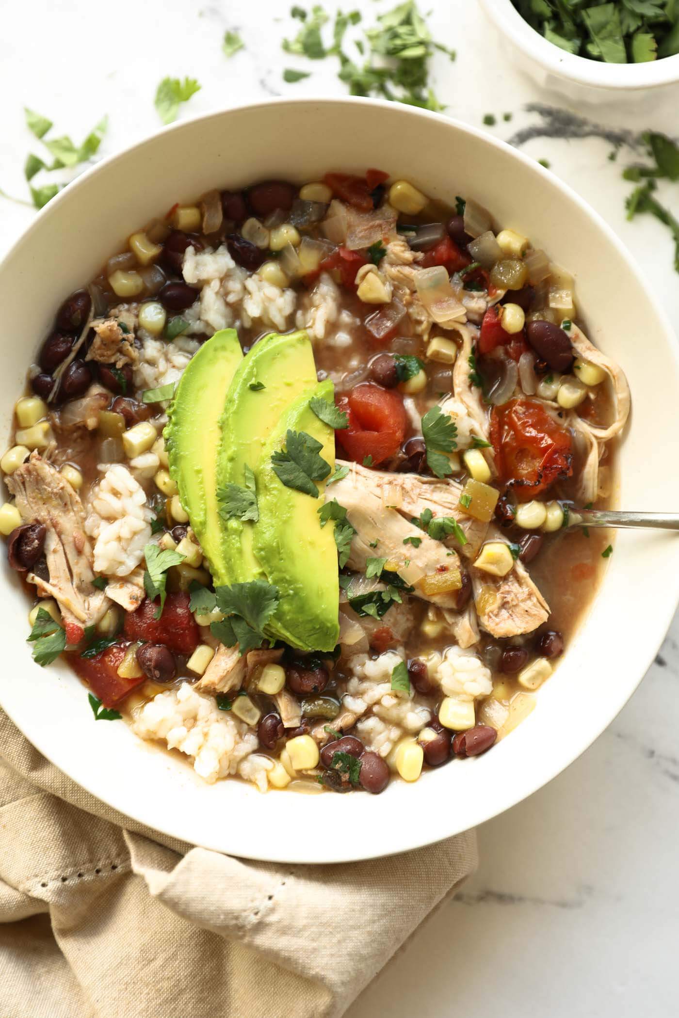 A bowl of chicken, rice, black bean, corn, and tomato soup topped with avocado slices and chopped cilantro, with a spoon and a napkin beside the bowl.