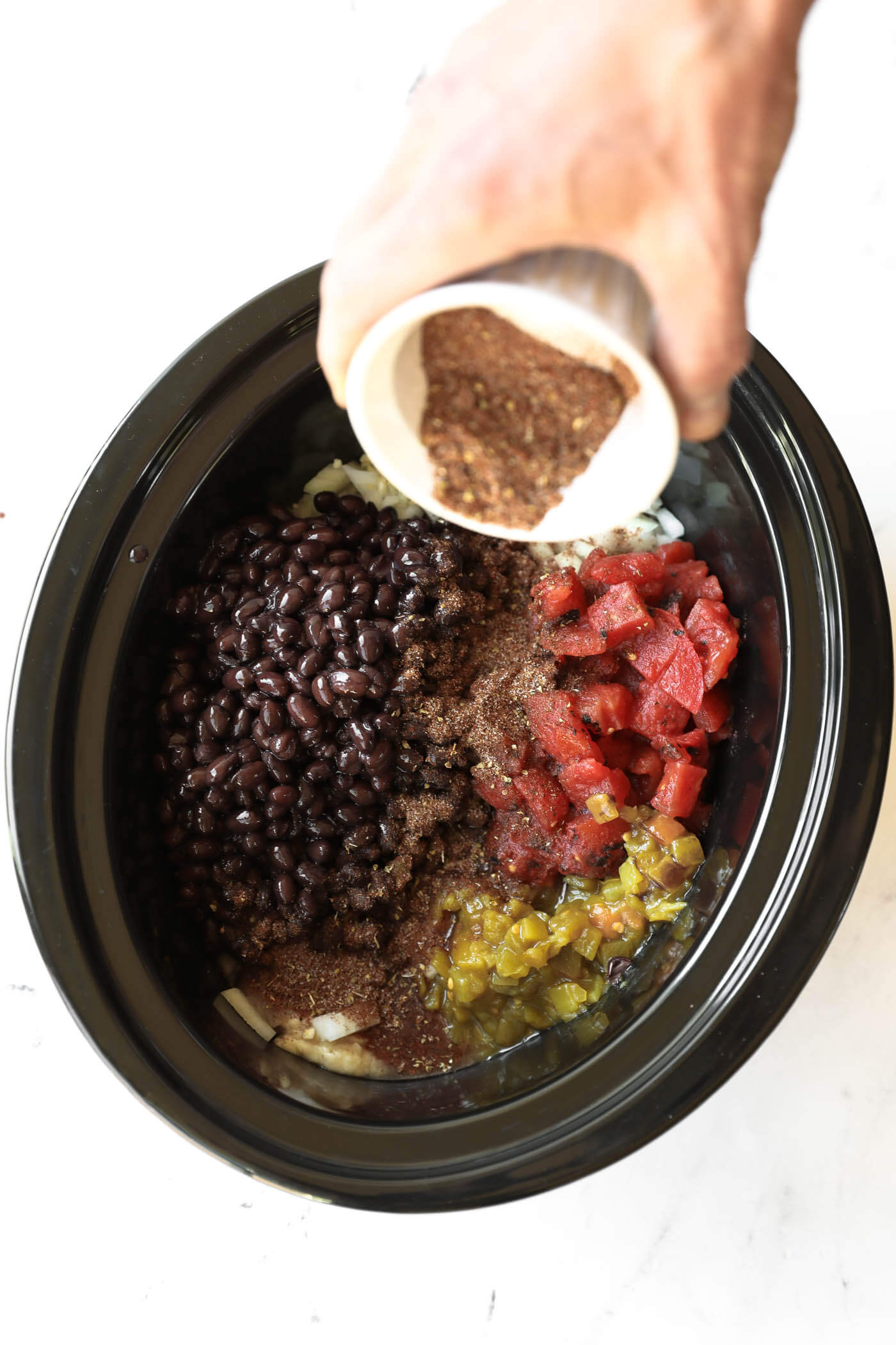 A hand pours spices into a slow cooker containing black beans, diced tomatoes, chopped onions, and green chilies.