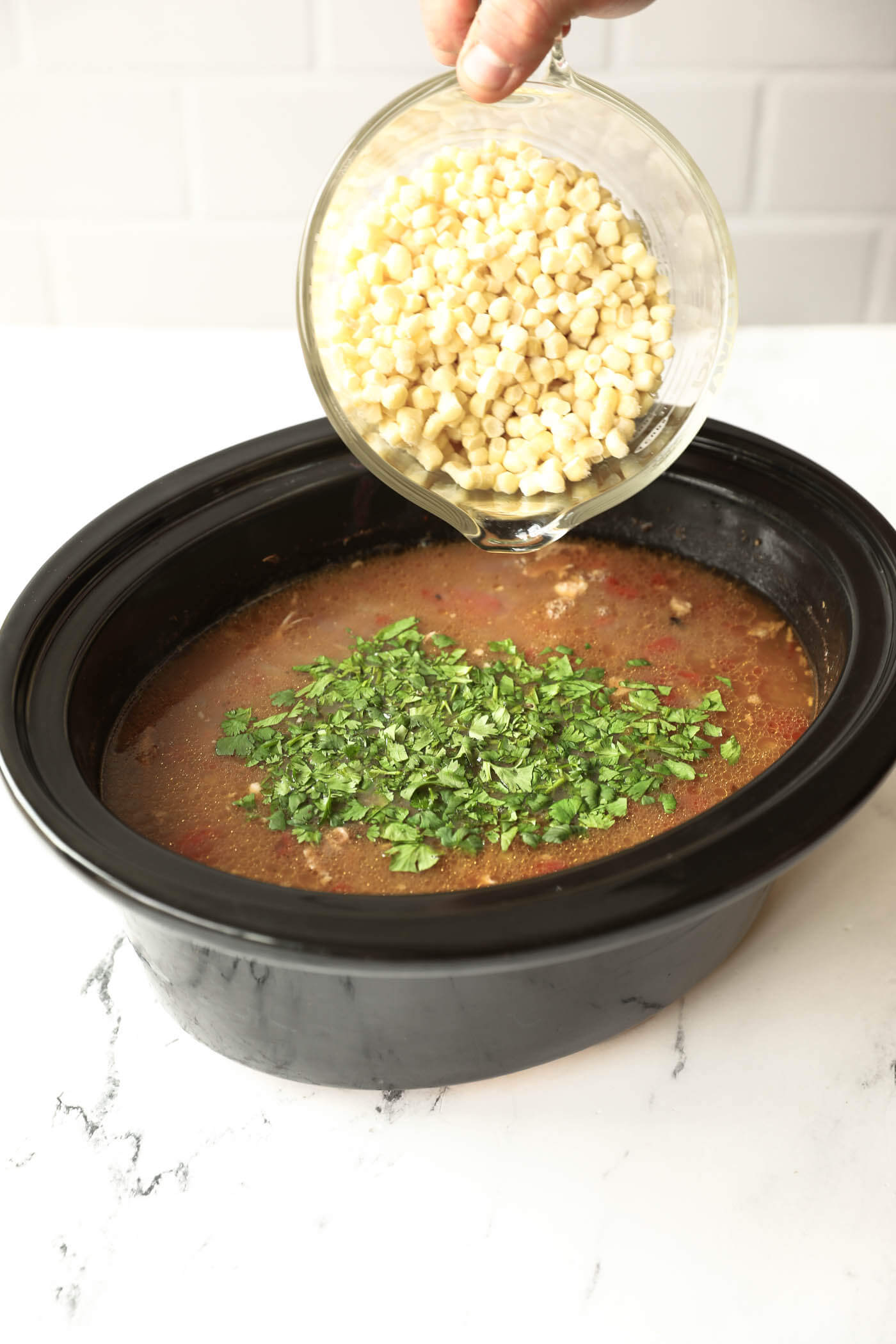 A hand pours a bowl of corn into a slow cooker filled with broth and chopped herbs on top.
