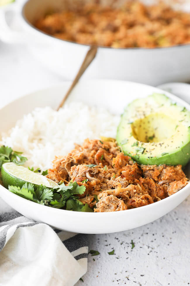 Close up angled image of crockpot mexican chicken in a bowl served with rice and avocado. Garnished with a lime wedge and fresh cilantro. 