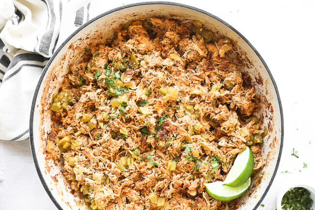 Horizontal image of crockpot mexican chicken in a skillet after being re-heated. Garnished with chopped cilantro and lime wedges. 