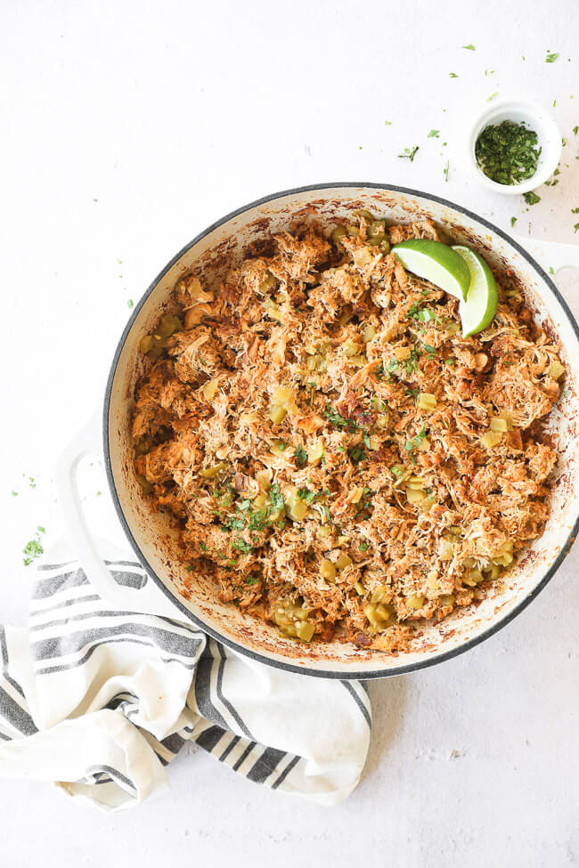 Vertical overhead image of crockpot mexican chicken in a skillet after re-heating. Garnished with cilantro and lime wedges. 