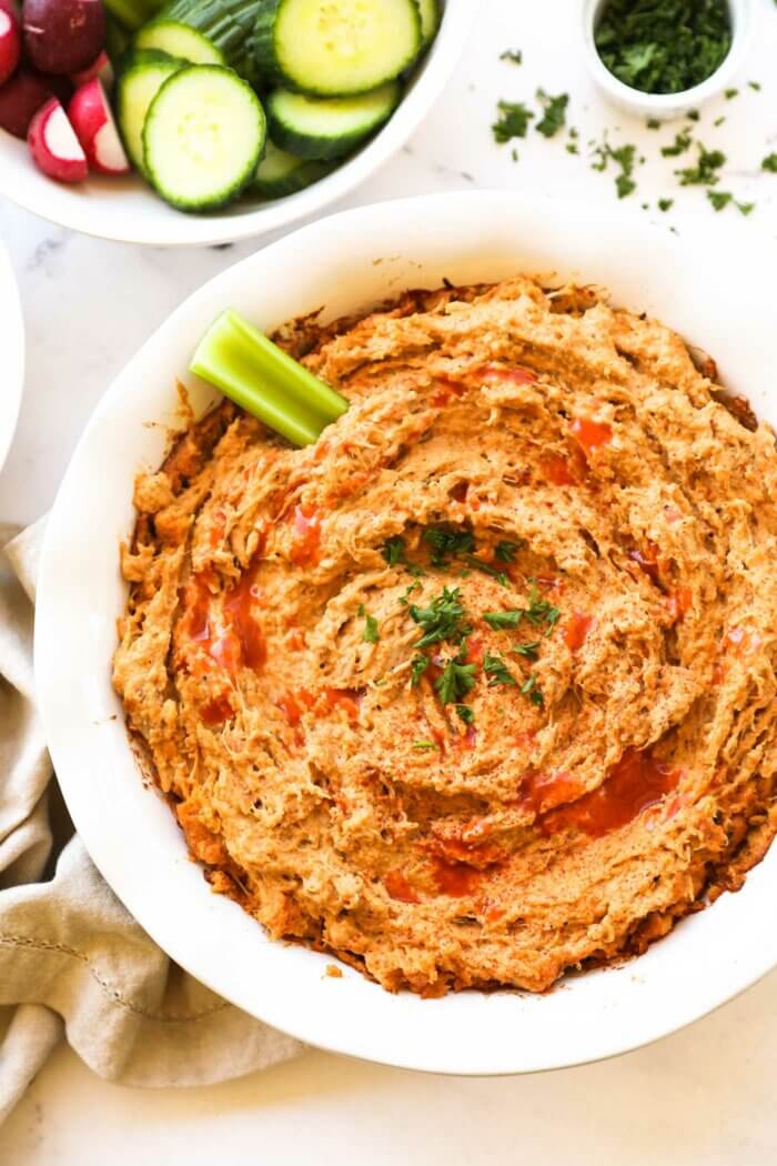 Overhead image of a round baking dish with buffalo chicken dip. A little extra hot sauce sprinkled on top, garnished with chopped parsley and a celery stick dug into the dip.