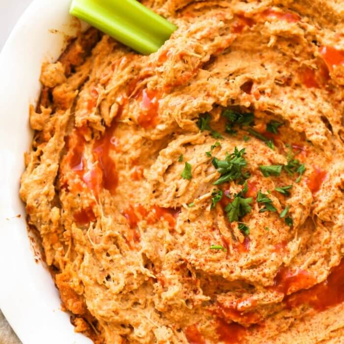 Close up overhead image of a round baking dish with buffalo chicken dip. A little extra hot sauce sprinkled on top, garnished with chopped parsley and a celery stick dug into the dip.