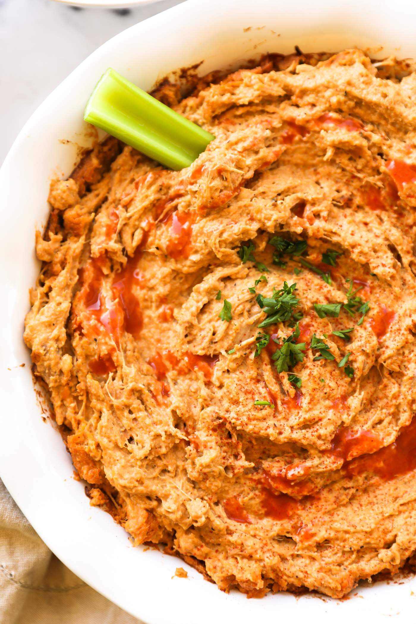 Close up overhead image of a round baking dish with buffalo chicken dip. A little extra hot sauce sprinkled on top, garnished with chopped parsley and a celery stick dug into the dip.