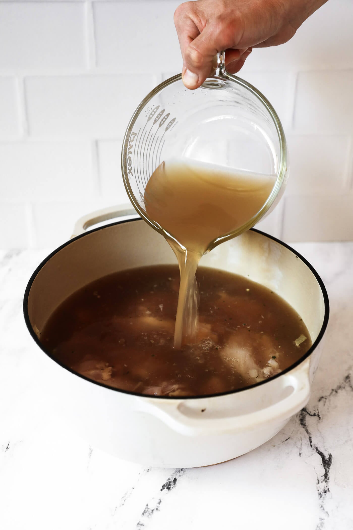 Pouring broth over chicken, veggies and seasonings in a dutch oven.