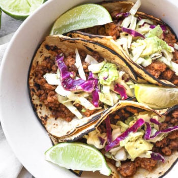 Three ground pork tacos with cabbage and avocado slaw in a bowl with lime wedges.