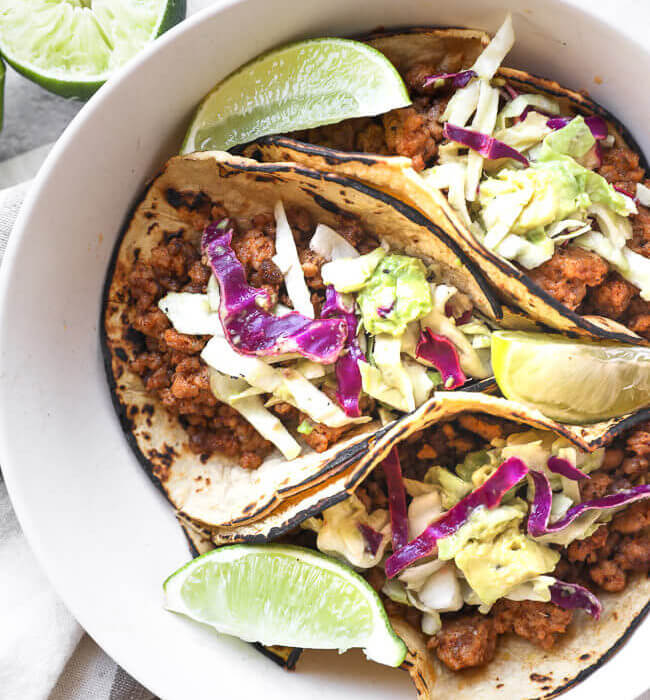 Three ground pork tacos with cabbage and avocado slaw in a bowl with lime wedges.