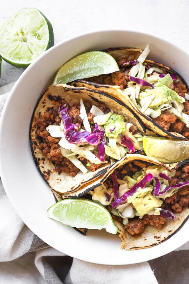 Overhead image of three ground pork tacos with cabbage and avocado slaw in a bowl with lime wedges.