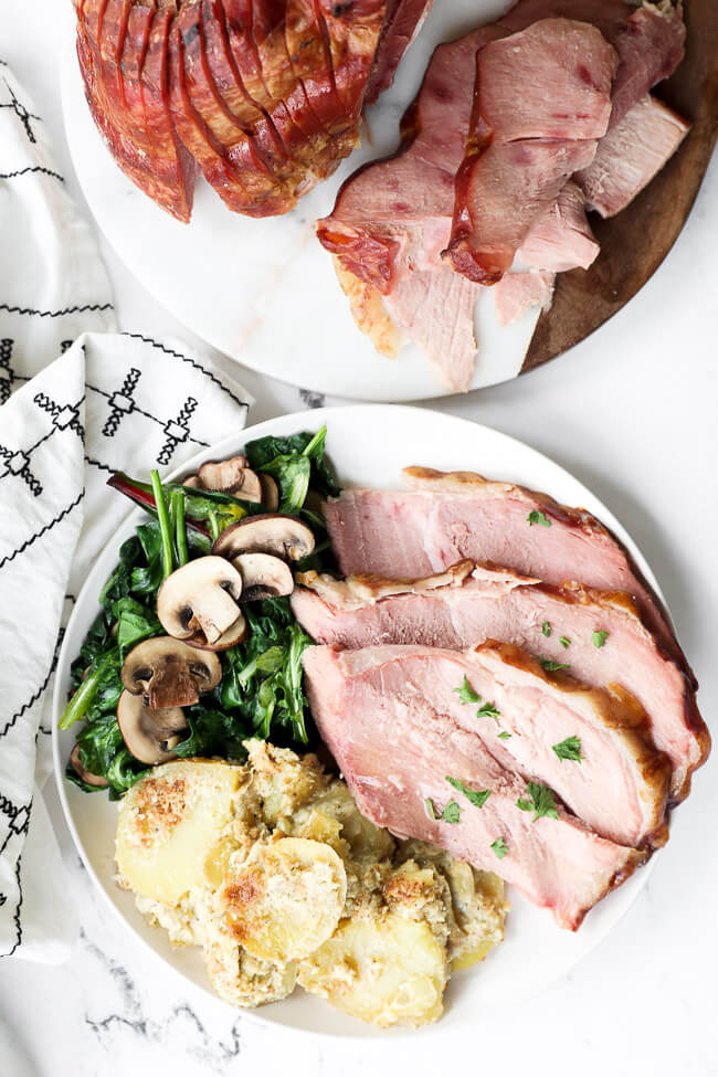 Overhead shot of sliced ham pieces on a plate with wilted greens and mushrooms and scalloped potatoes