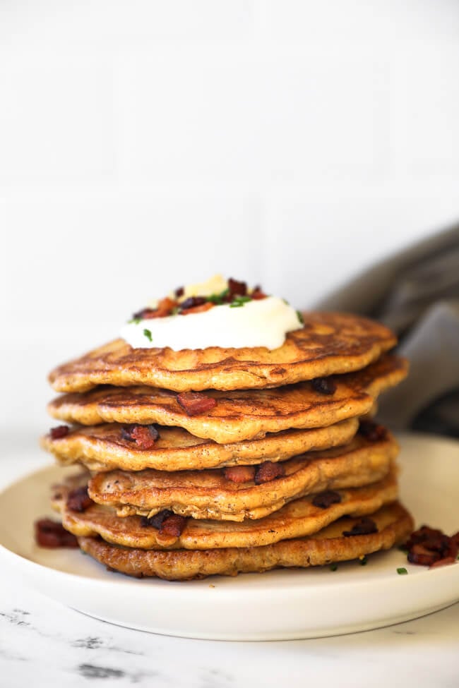 Stack of bacon pancakes on a plate with toppings including sour cream, bacon, chives and cheese. 