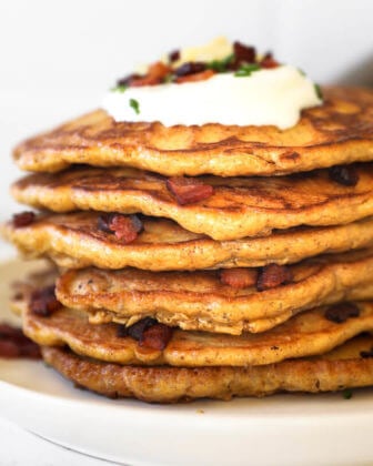 stack of bacon pancakes on a plate with sour cream, chives, more bacon and shredded cheese on top.