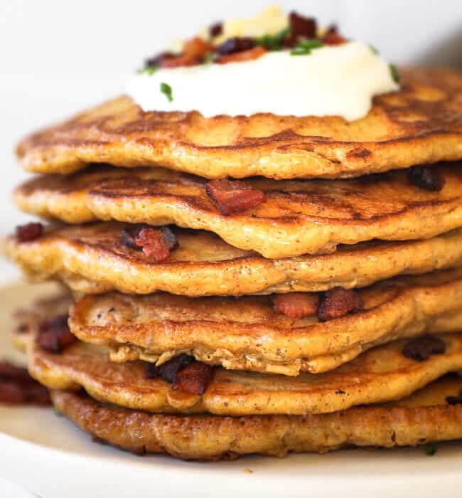 stack of bacon pancakes on a plate with sour cream, chives, more bacon and shredded cheese on top.