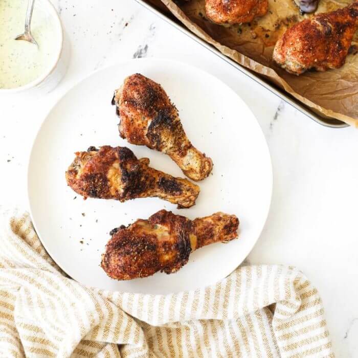 3 Baked chicken drumsticks on a white plate with sheet pan and sauce next to plate