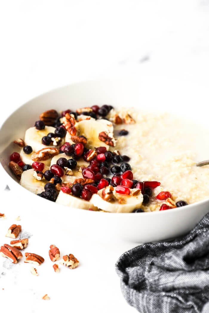 Angled image of a bowl of creamy millet porridge with coconut milk, bananas, berries, pomegranate seeds and chopped pecans.