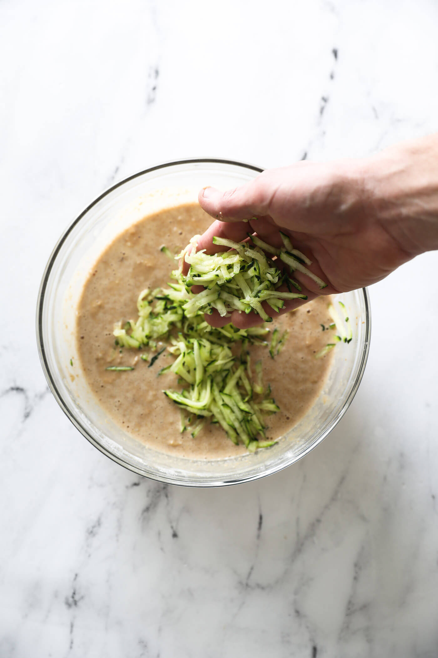 A hand adding the shredded zucchini to the batter in a mixing bowl.