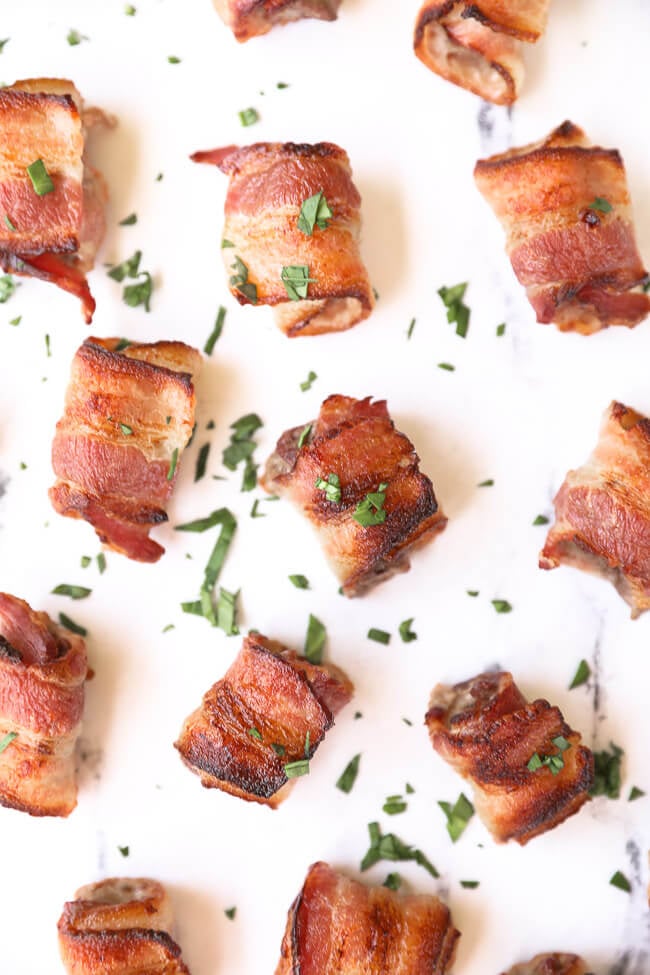 Overhead image of bacon wrapped steak bites spread out on marble with chopped parsley sprinkled about. 