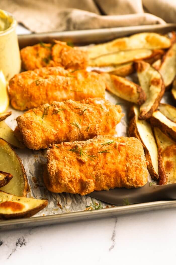 Angled image of four pieces of battered and baked cod with potato wedges on a sheet pan. The piece of fish closest to the camera is being lifted with a spatula.