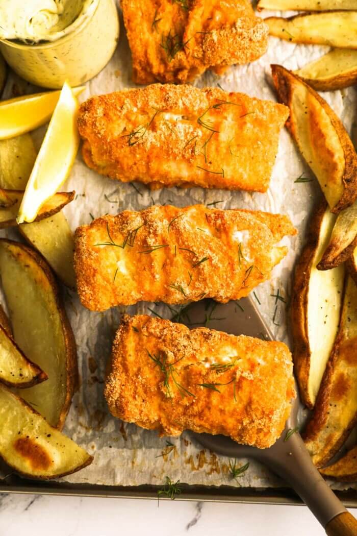 Close up of gluten free fish and chips on a sheet pan. One piece of fish is being lifted with a spatula.