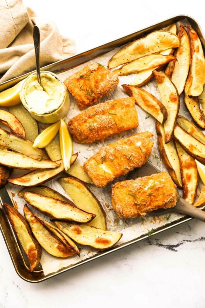 Overhead image of a sheet pan with baked fish and chips. The battered fish pieces are in the middle of the sheet pan with baked potato wedges on the top and bottom of the pan. A jar of homemade tartar sauce is also on the pan with lemon wedges to garnish.