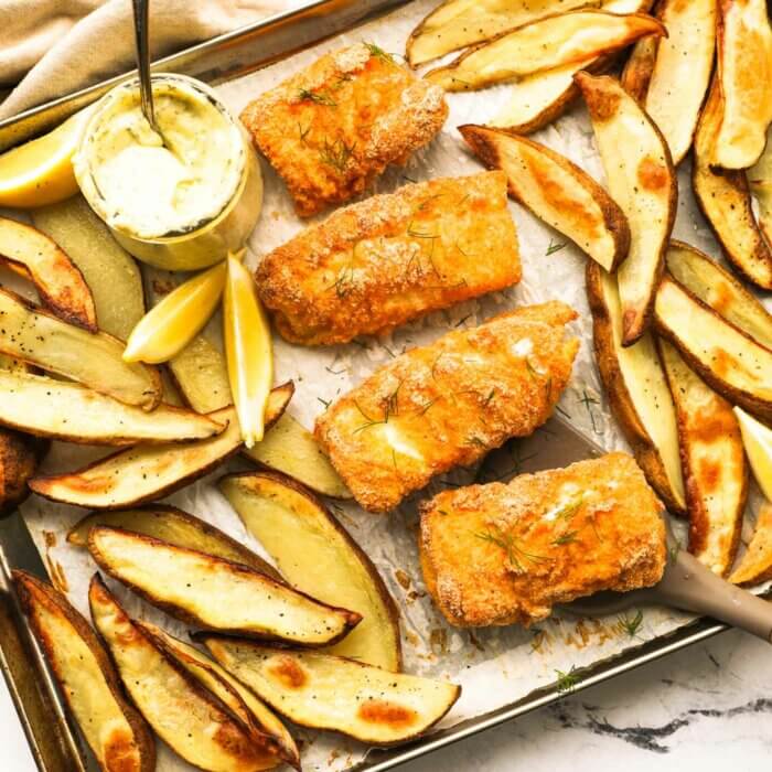 Overhead image of a sheet pan with baked fish and chips. The battered fish pieces are in the middle of the sheet pan with baked potato wedges on the top and bottom of the pan. A jar of homemade tartar sauce is also on the pan with lemon wedges to garnish.
