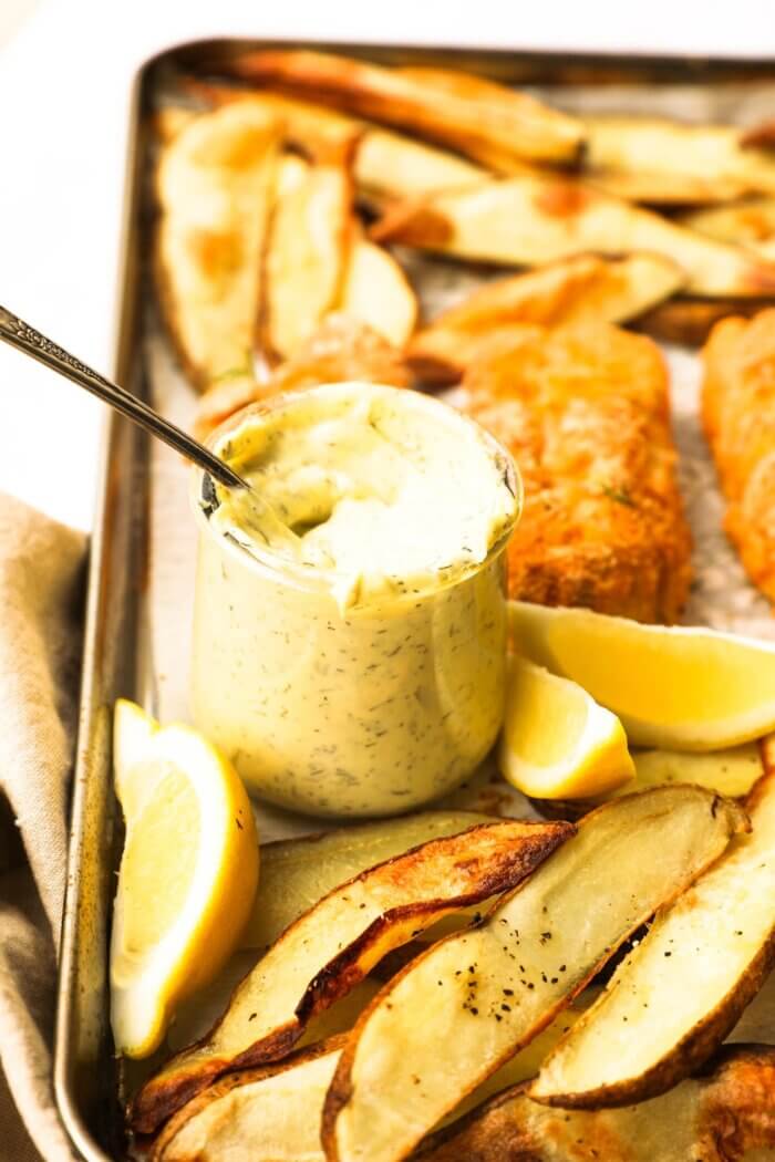 Homemade tartar sauce in a jar with a spoon on the sheet pan with gluten free fish and chips.