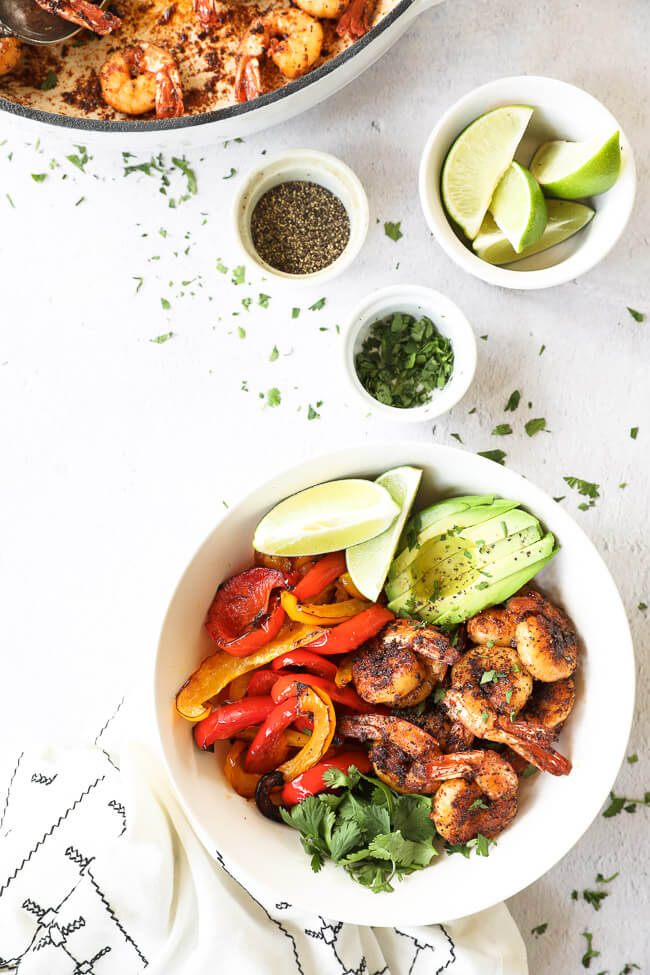 Overhead vertical image of shrimp in  a bowl with roasted red and yellow peppers, avocado, lime wedges and fresh cilantro. Skillet with extra shrimp on the side as well as black pepper, cilantro and lime wedges. 
