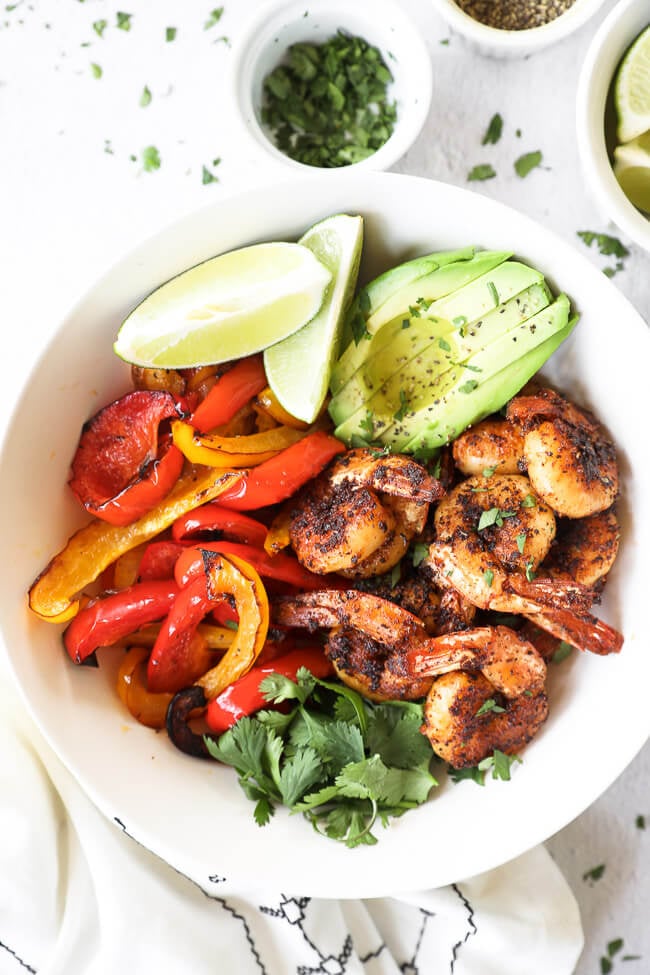 Vertical close up image of blackened shrimp served in a bowl with roasted red and yellow peppers, sliced avocado, lime wedges and fresh cilantro. 