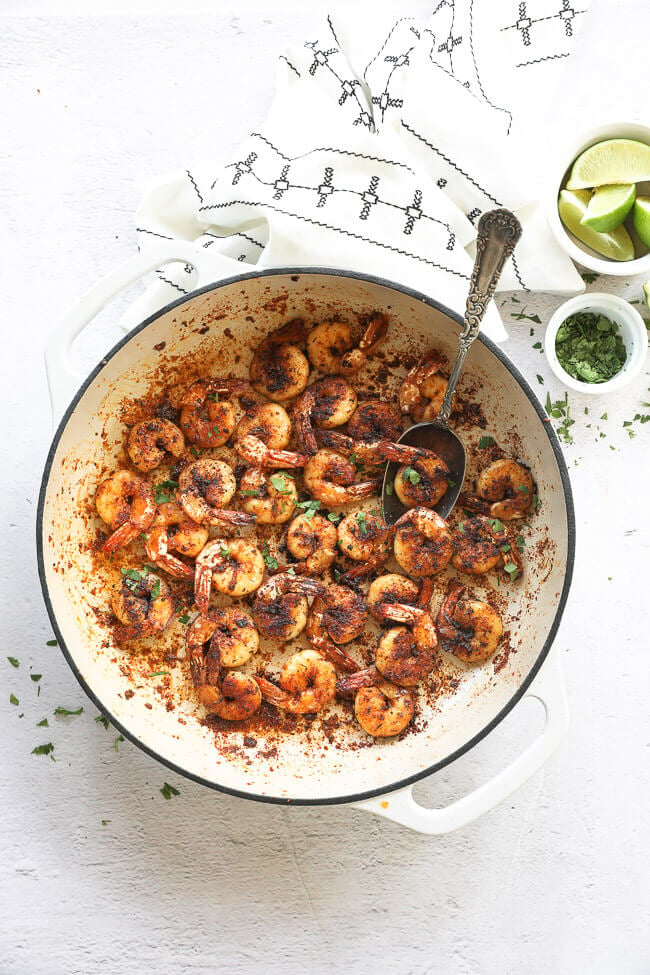 Overhead vertical image of blackened shrimp in a skillet with fresh chopped cilantro sprinkled on top and a spoon like it's scooping shrimp out. 