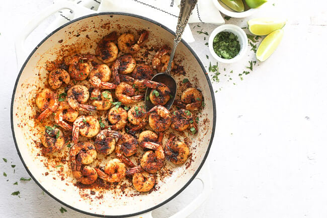 Horizontal overhead image of blackened shrimp in a skillet with chopped cilantro sprinkled on top and a spoon like it's scooping some shrimp out. 