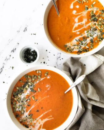 Two bowls of butternut squash and roasted red pepper soup with coconut milk drizzled on top with pumpkin seeds and chopped oregano.