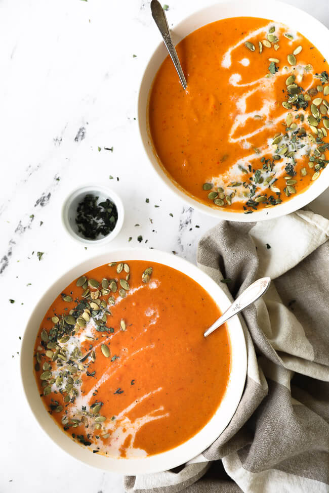 Two bowls of butternut squash and roasted red pepper soup with coconut milk drizzled on top with pumpkin seeds and chopped oregano.