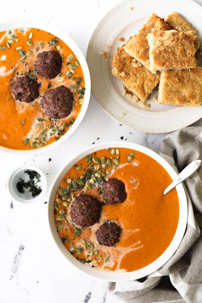 Two bowls of butternut squash and roasted red pepper soup with coconut milk drizzled on top with pumpkin seeds and chopped oregano. Served with meatballs and a side of paleo cornbread. 