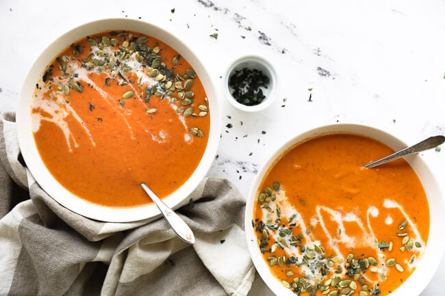 Two bowls of butternut squash and roasted red pepper soup with coconut milk drizzled on top with pumpkin seeds and chopped oregano. 
